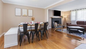 Uma sala de estar com lareira, um espaço de jantar com mesa e cadeiras, e uma televisão fixada na parede.