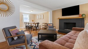 Uma sala de estar com lareira, televisão na parede e um espaço de jantar com cadeiras e mesa.