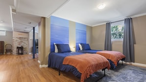 Quarto com duas camas, parede de destaque em tijolo, ampla janela com cortinas e piso de madeira.