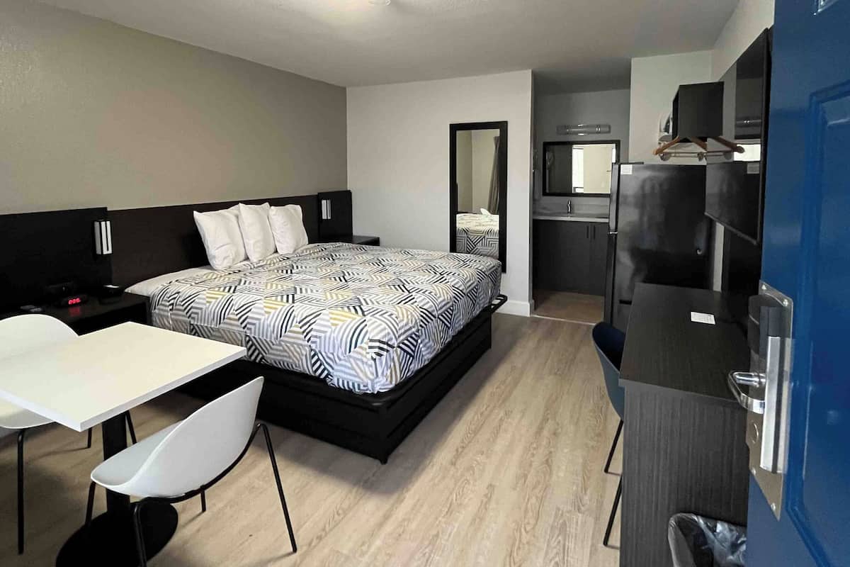 A compact hotel room with a bed, a small desk, a chair, a kitchenette, and a mirror.