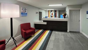 A reception area with a desk, a chair, a lamp, a striped carpet, and a door. - Studio 6 Conroe, TX (Conroe)