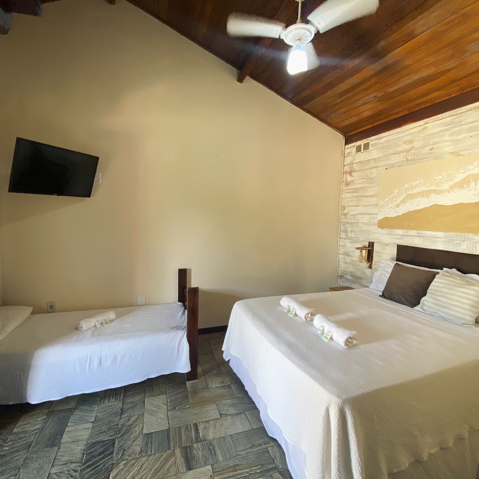 A hotel room with two beds, a ceiling fan, a mounted TV, and a stone-patterned floor.