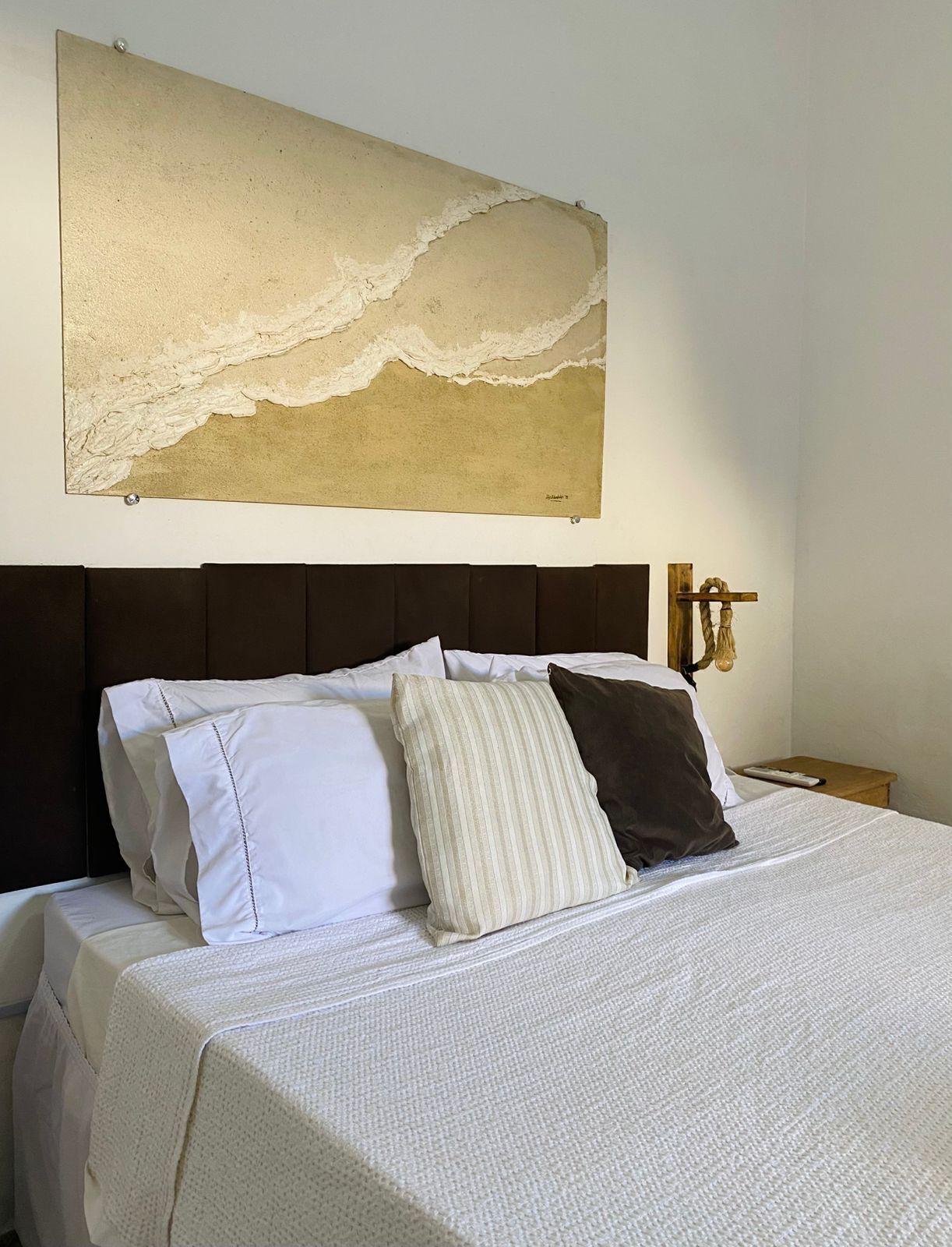 A neatly made bed with white and striped pillows, a textured white bedspread, and a large abstract painting above it.