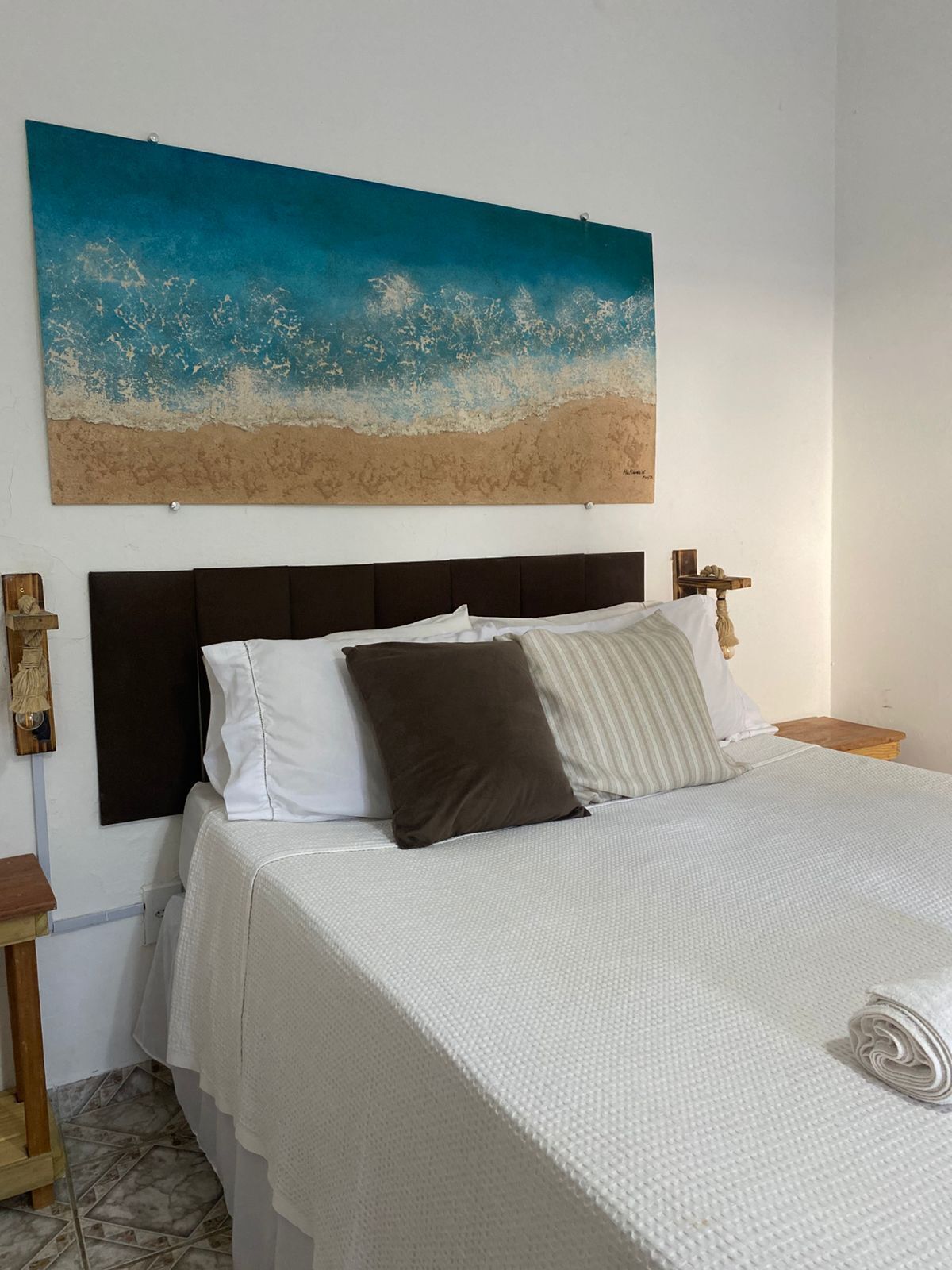 Una cama con una colcha blanca, un cabecero y un cuadro de una playa sobre él.