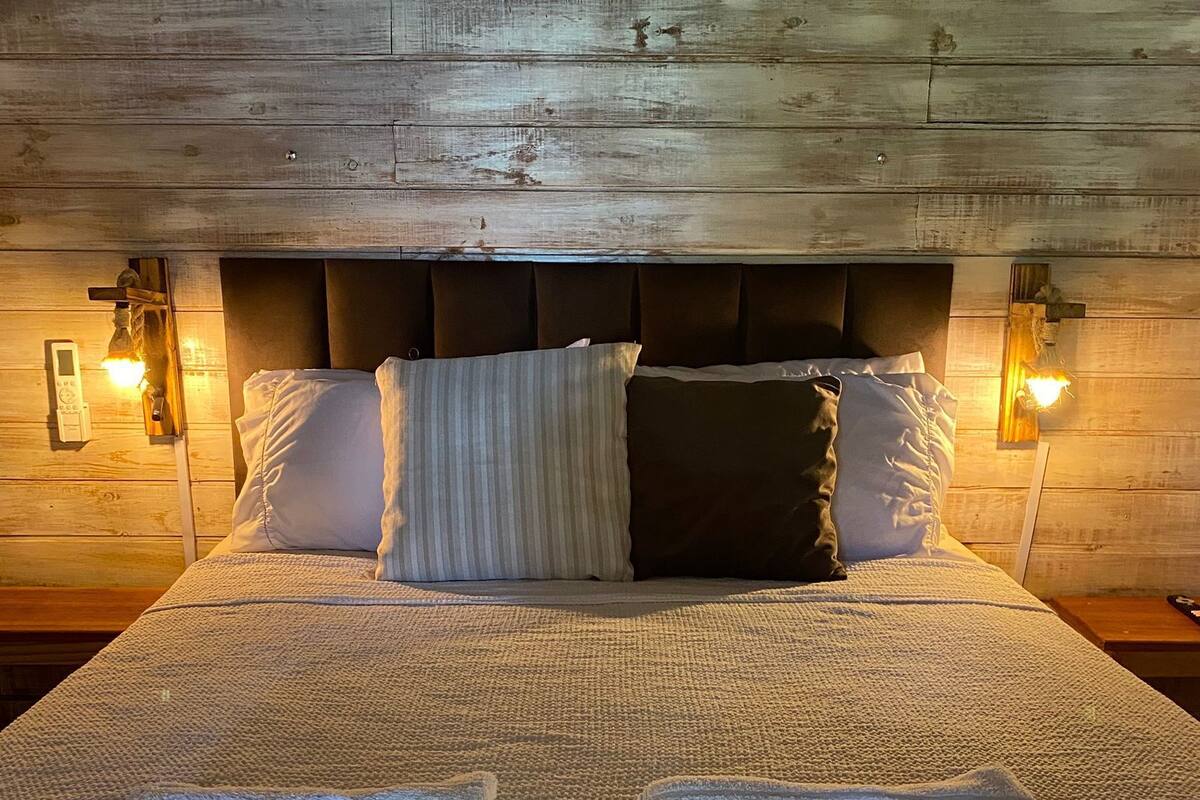 A neatly made bed with pillows and two towels, wooden headboard, and wall-mounted lights.