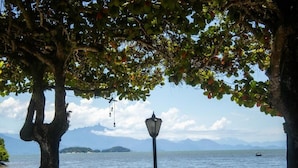 Vista al mar con una mesa y sillas bajo un árbol, un poste de luz y un barco a lo lejos en el agua.