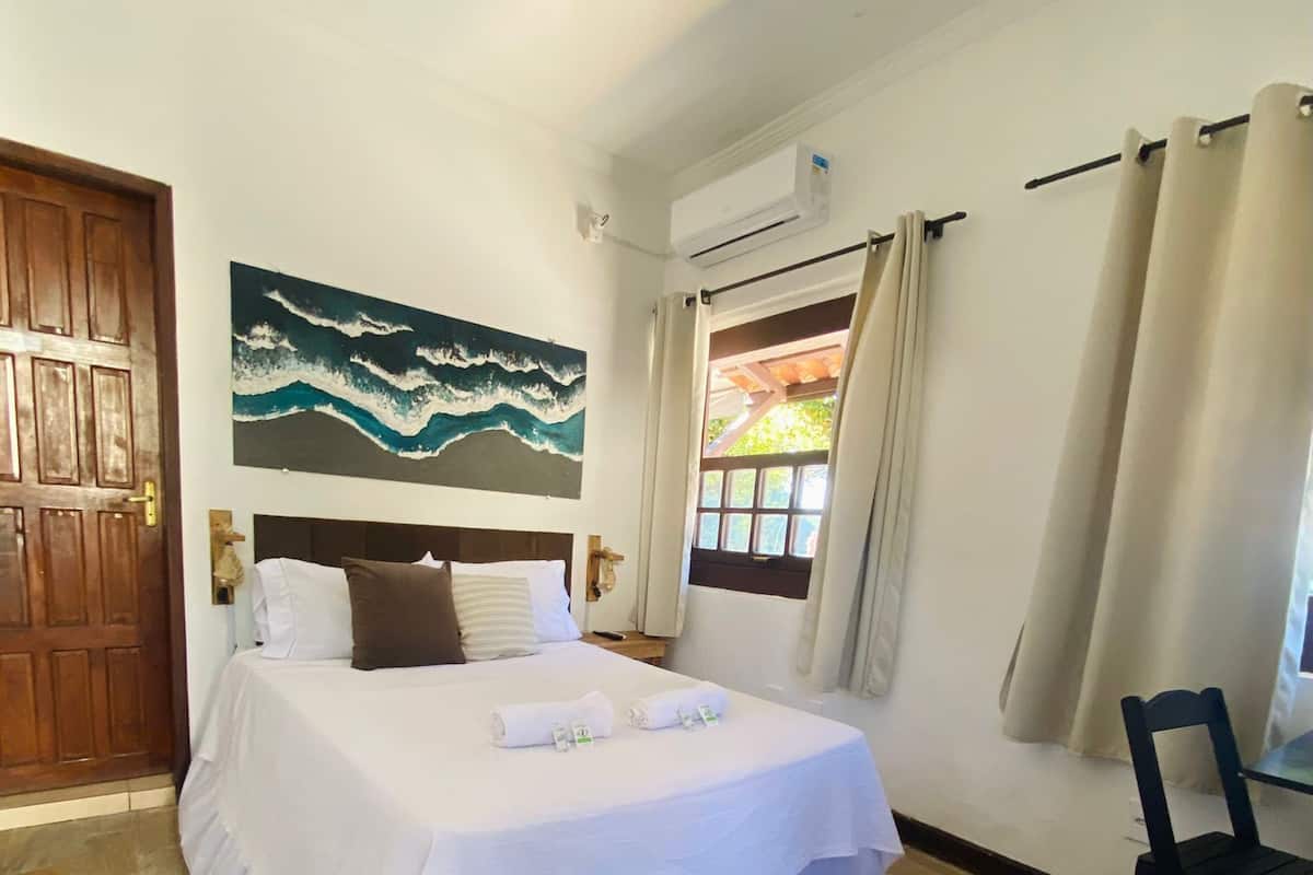 A hotel room with a bed, a wooden door, a painting, a window with curtains, and a chair.