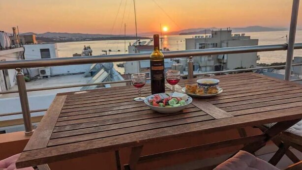 A wooden table with a bottle, glasses, and food on a balcony overlooking a sunset. - 5th-Floor Cozy Sea View, 2-Min Beach Walk (Chania)