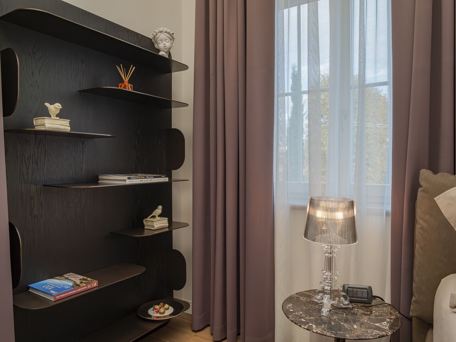 Una sala de estar moderna con una estantería de madera oscura, una mesita redonda con una lámpara, y vistas al exterior a través de cortinas traslúcidas.