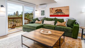 Un salon moderne avec un canapé d’angle vert, une table basse en bois et une grande fenêtre donnant sur une terrasse.