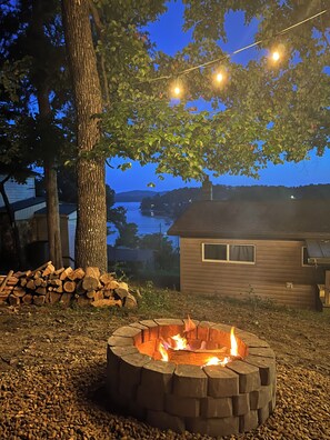 A backyard with a fire pit, a stack of firewood, a tree, and a house with a view of a body of water. - Beautiful 2 bedroom cabin with amazing lake views (Bean Station)