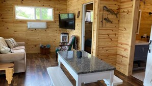 Interior acolhedor de uma cabana, com decoração em madeira, televisão na parede, um sofá, uma mesa de jantar e um banco.