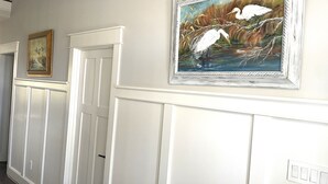 A hallway with white wainscoting and two framed artworks on the wall. - Custom 4-bedroom home, boat slip, fenced backyard, pool & paved bike path. (Cape Charles)
