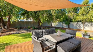 An outdoor patio with a table and chairs, surrounded by a wooden deck and a large shade sail.