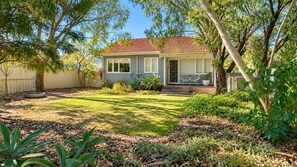 A residential property with a well-maintained lawn, a front porch with two chairs, and a variety of plants and trees surrounding the house. - Barefoot Beach Cottage (Geographe)