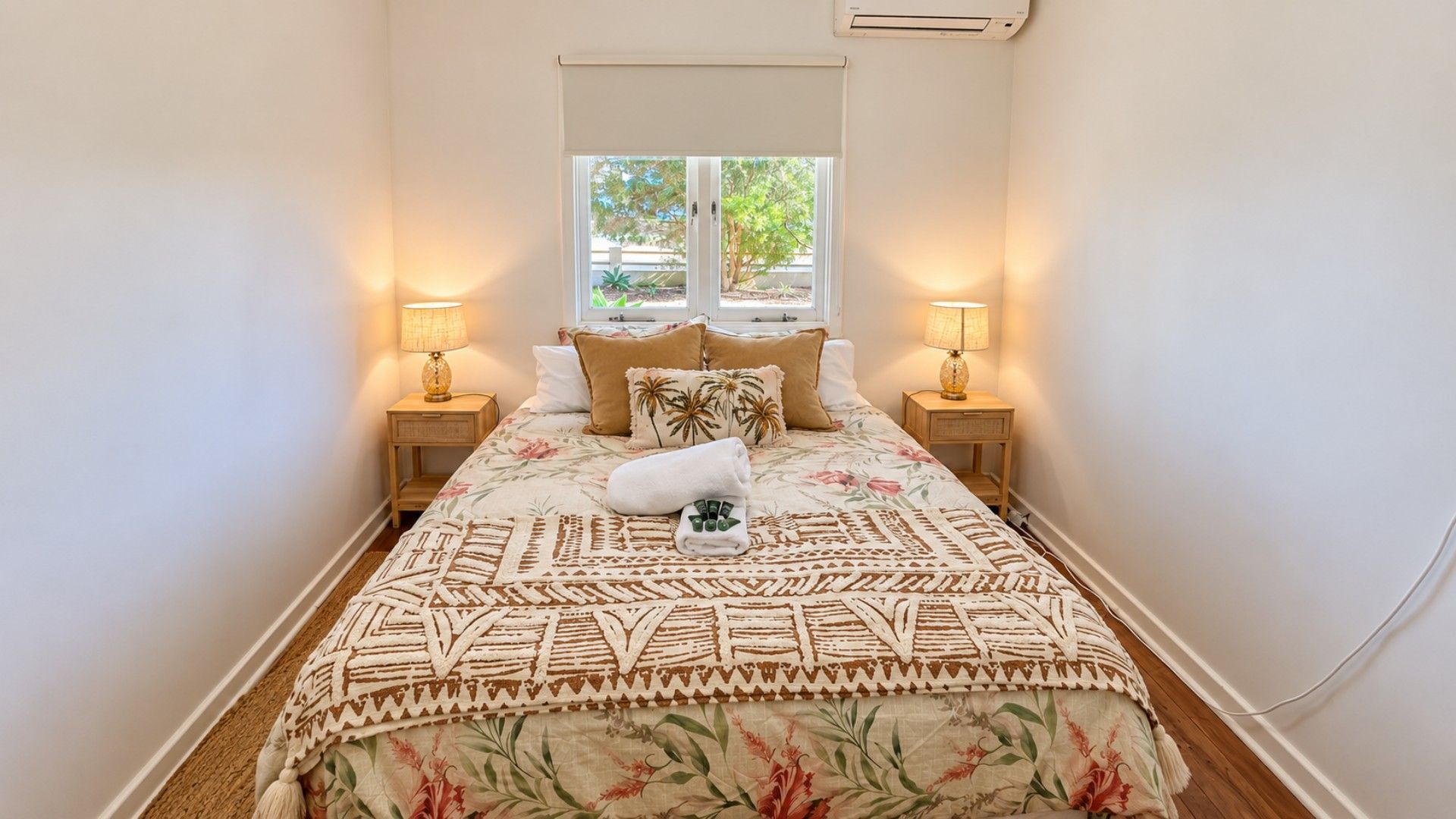 A narrow hotel room with a bed, two bedside lamps, a window with a view of greenery, and a wall-mounted air conditioning unit.