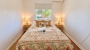 A narrow hotel room with a bed, two bedside lamps, a window with a view of greenery, and a wall-mounted air conditioning unit. - Barefoot Beach Cottage (Geographe)