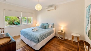 A bedroom with a bed, bedside tables, a chair, a window with a view of greenery, and an air conditioning unit.
