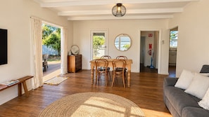 A living room with a sofa, a dining table with chairs, a TV, and a large mirror. - Barefoot Beach Cottage (Geographe)