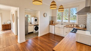 A modern kitchen with wooden flooring, white cabinetry, a built-in oven, and a large window providing natural light. - Barefoot Beach Cottage (Geographe)