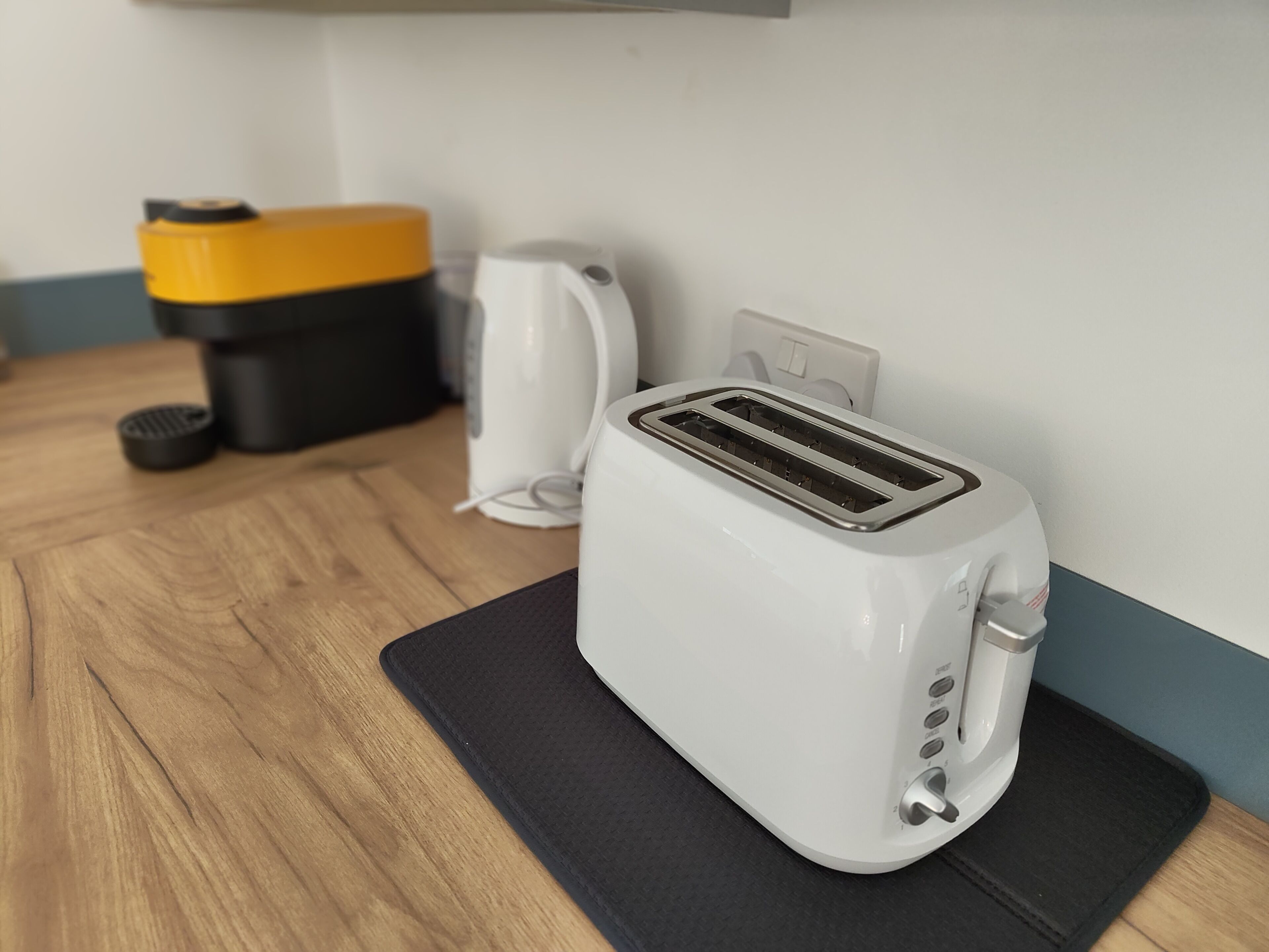 A white toaster on a black mat, a yellow and black container, and a white kettle on a wooden table.