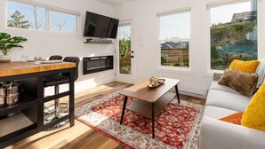 A modern living room with a dining area, a flat-screen TV mounted on the wall, and large windows offering views of a garden and a building. - New Garden Studio I ProClean I Skylight I Rain Shower I Nespresso I Free Parking (Colwood)