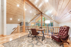 A spacious loft bedroom with a wooden ceiling, a large window, a wooden desk, a leather chair, and a patterned rug.