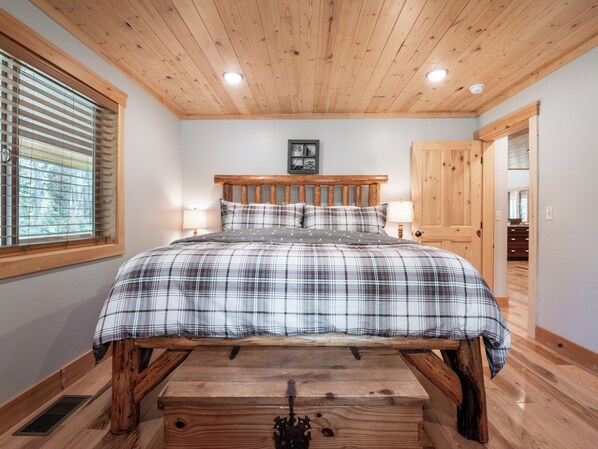 A bedroom with a wooden headboard, a plaid bedspread, two bedside lamps, and a window with blinds.