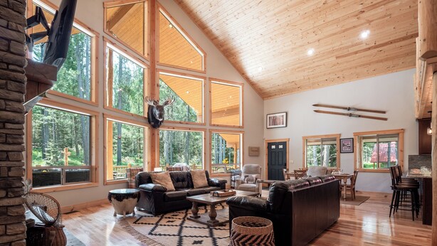 A spacious living room with a large fireplace, wooden flooring, and large windows offering a forest view. - Hot Tub, Firepit, Large Deck, Game Room, Secluded Mtn Views, 20 min to Park | Yellowstone Escape (Island Park)