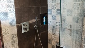 Een douchecabine met glazen wanden, een handdouche, bedieningspaneel en een betegelde muur met een geometrisch patroon.
