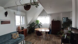 Een ruime woonkamer met hoge plafonds, een eettafel met stoelen, een bank en diverse potplanten.