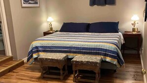 A bedroom with a wooden floor, a bed with a striped blanket, two bedside tables with lamps, a dark curtain, and a framed picture on the wall.