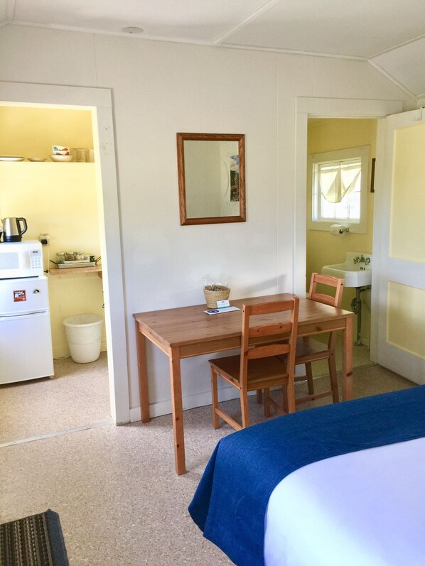 A compact room with a bed, a wooden desk, a chair, a small kitchenette, and a mirror on the wall.
