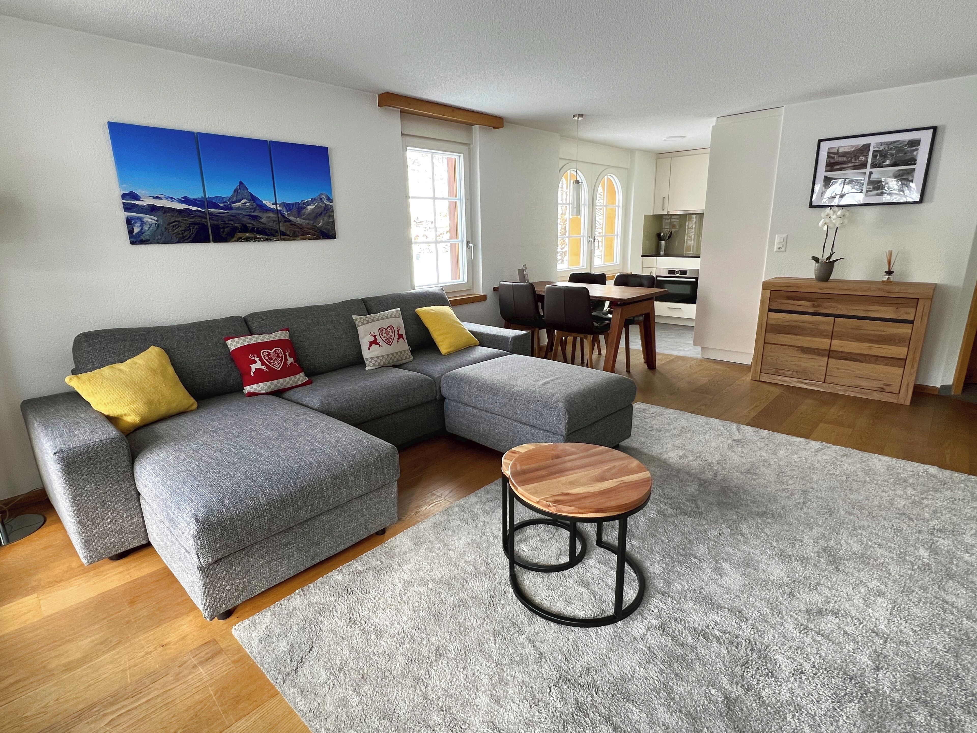 Een moderne woonkamer met een grijze hoekbank, een rond houten bijzettafeltje en een eethoek met een houten tafel en stoelen.