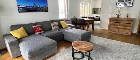 Een moderne woonkamer met een grijze hoekbank, een rond houten bijzettafeltje en een eethoek met een houten tafel en stoelen.