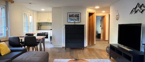 Uma sala de estar moderna com um sofá, dois banquinhos de madeira, uma TV e um espaço de refeições com cadeiras e uma mesa.