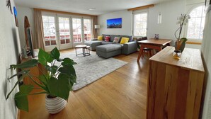 A spacious living room with a grey sofa, a wooden dining table, and a large window. - Haus Vira (Zermatt)