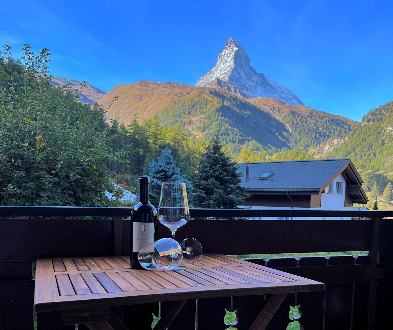 A wooden table with a bottle and glasses, overlooking a mountain with a distinctive peak.