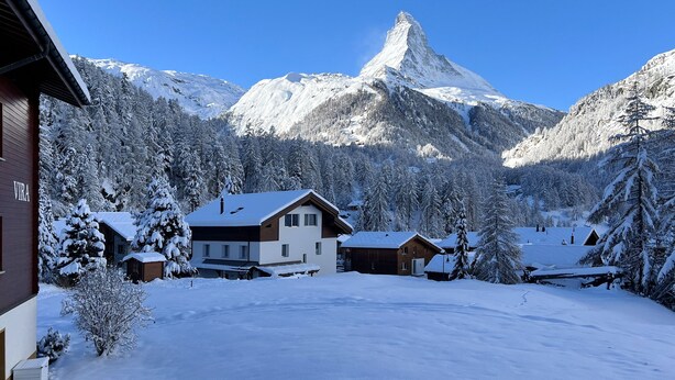 A snow-covered mountain with a prominent peak, surrounded by residential buildings with snow-covered roofs and trees. - Haus Vira (Zermatt)