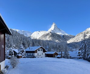 A snow-covered mountain with a prominent peak, surrounded by residential buildings with snow-covered roofs and trees. - Haus Vira (Zermatt)