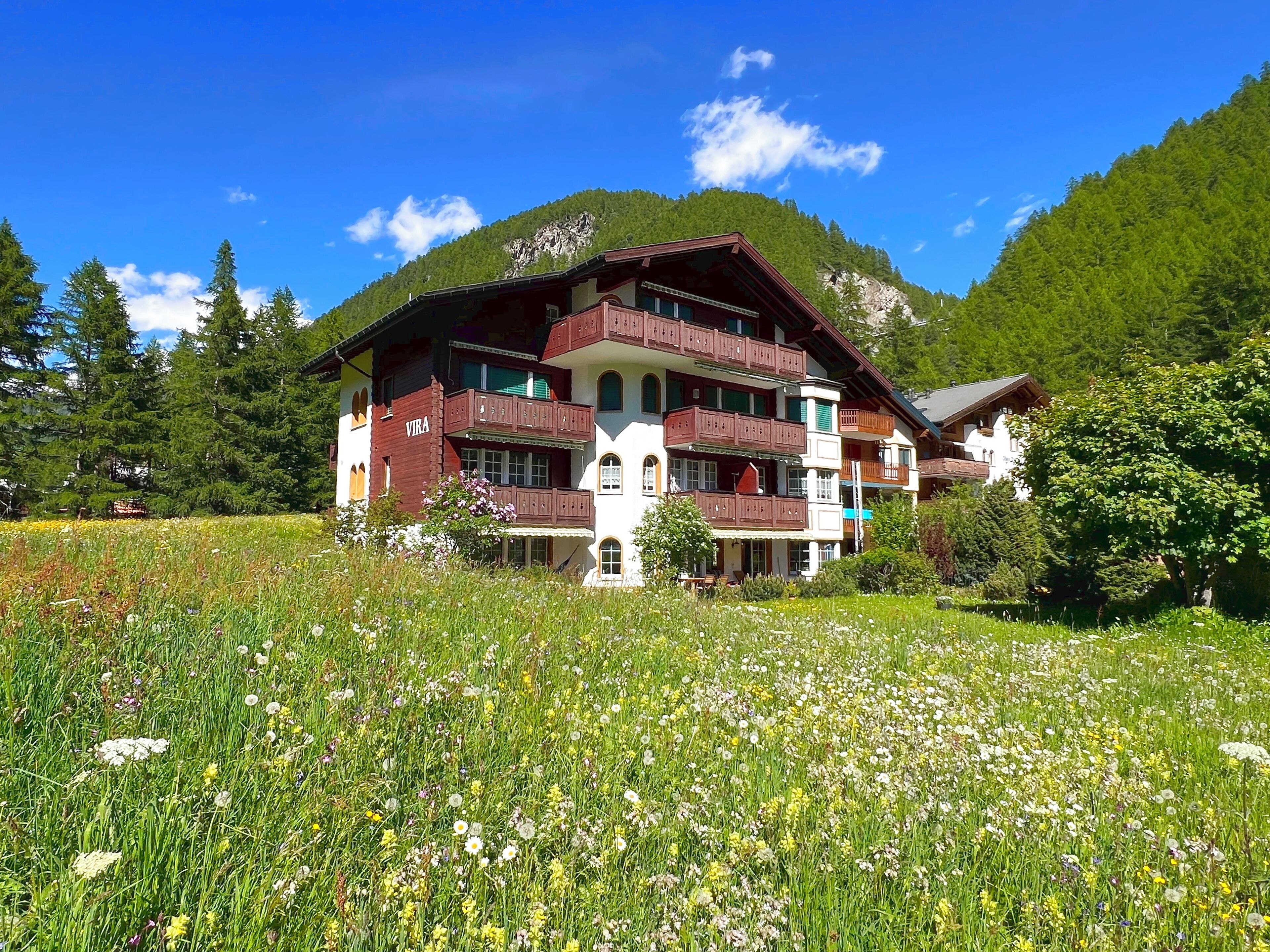 A multi-story building with balconies, surrounded by a lush meadow and mountains.