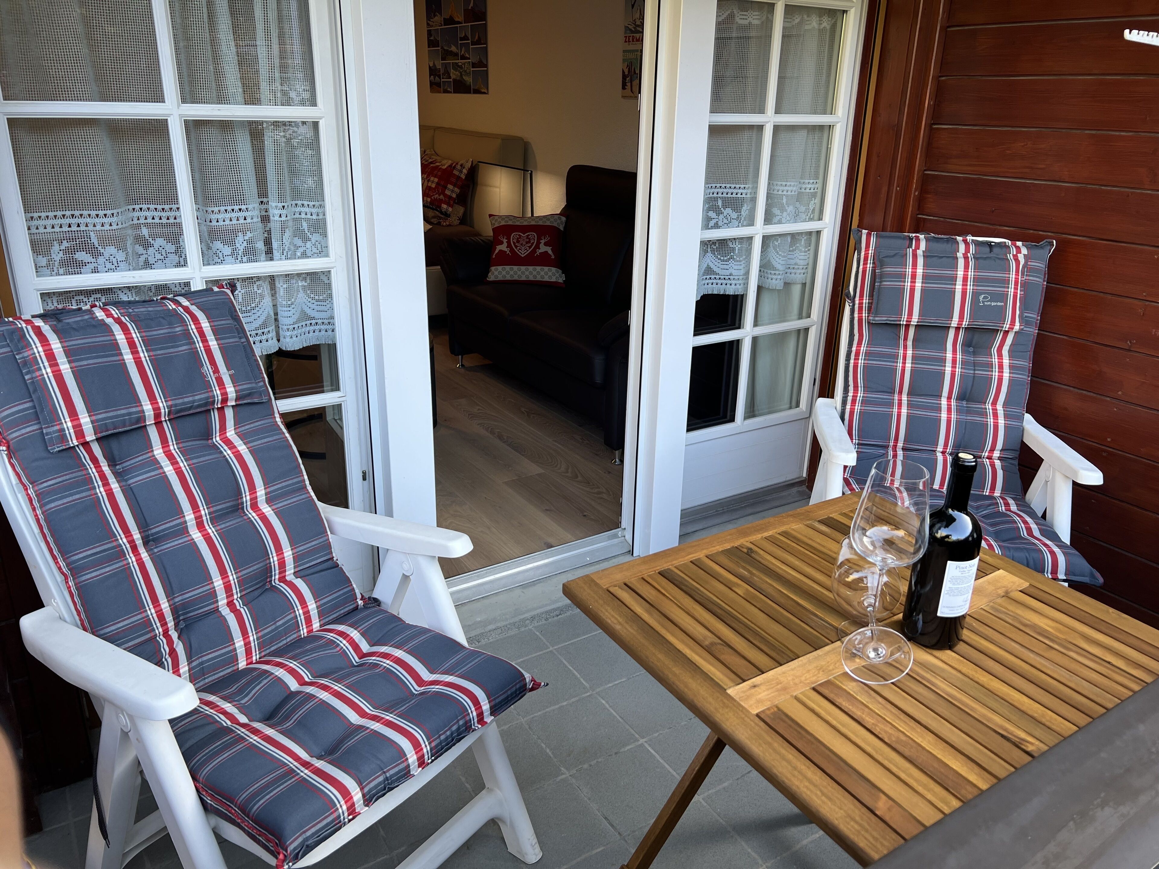 Eine Terrassenfläche mit zwei gestreiften Stühlen und einem Holztisch, einer Weinflasche und einem Glas auf dem Tisch.