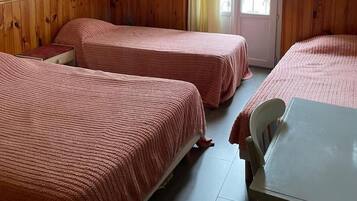 A room with two beds, a table, a chair, a picture on the wall, and a window with curtains.