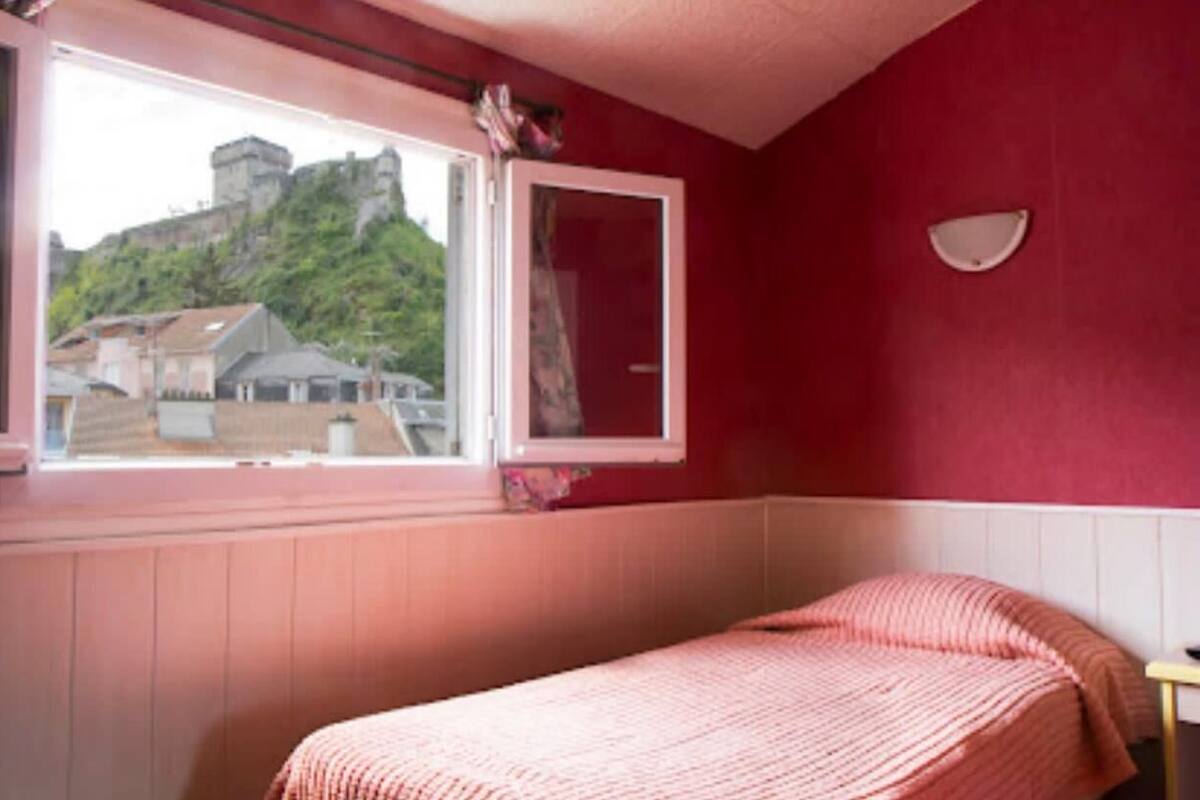 Quarto com cama, janela com vista para um edifício e vegetação, e luminária fixada na parede.
