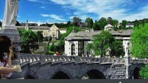 A stone bridge with a statue on the left, a clear blue sky, and buildings in the background. - N.D. Auxiliatrice (Lourdes)