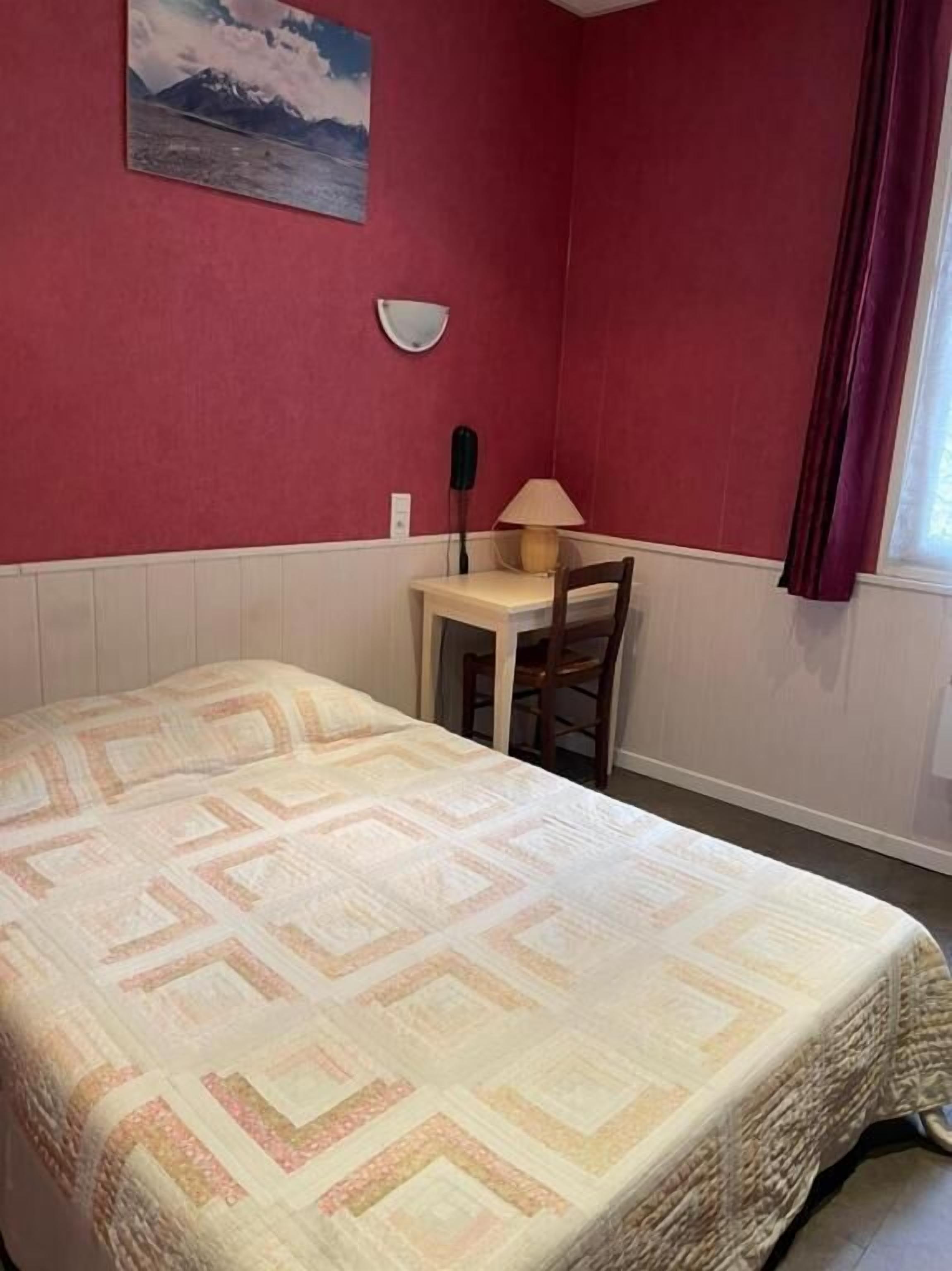 A room with a bed, a desk, a chair, a lamp, and a painting on the wall.