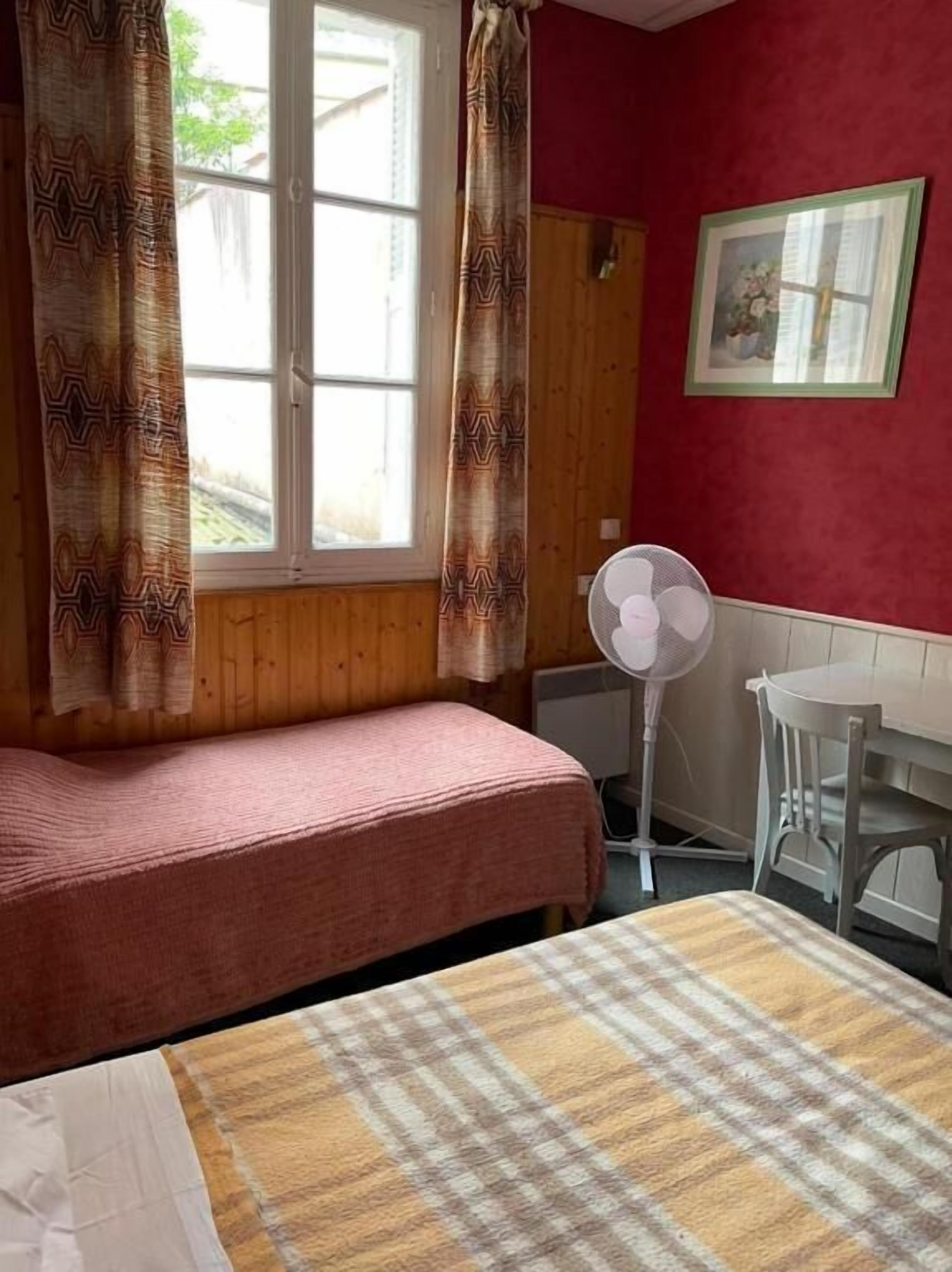 Una habitación con una cama, una mesa, una silla, un ventilador y una ventana con cortinas.