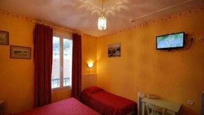 A room with two beds, a desk, a television, and a window with curtains. - N.D. Auxiliatrice (Lourdes)