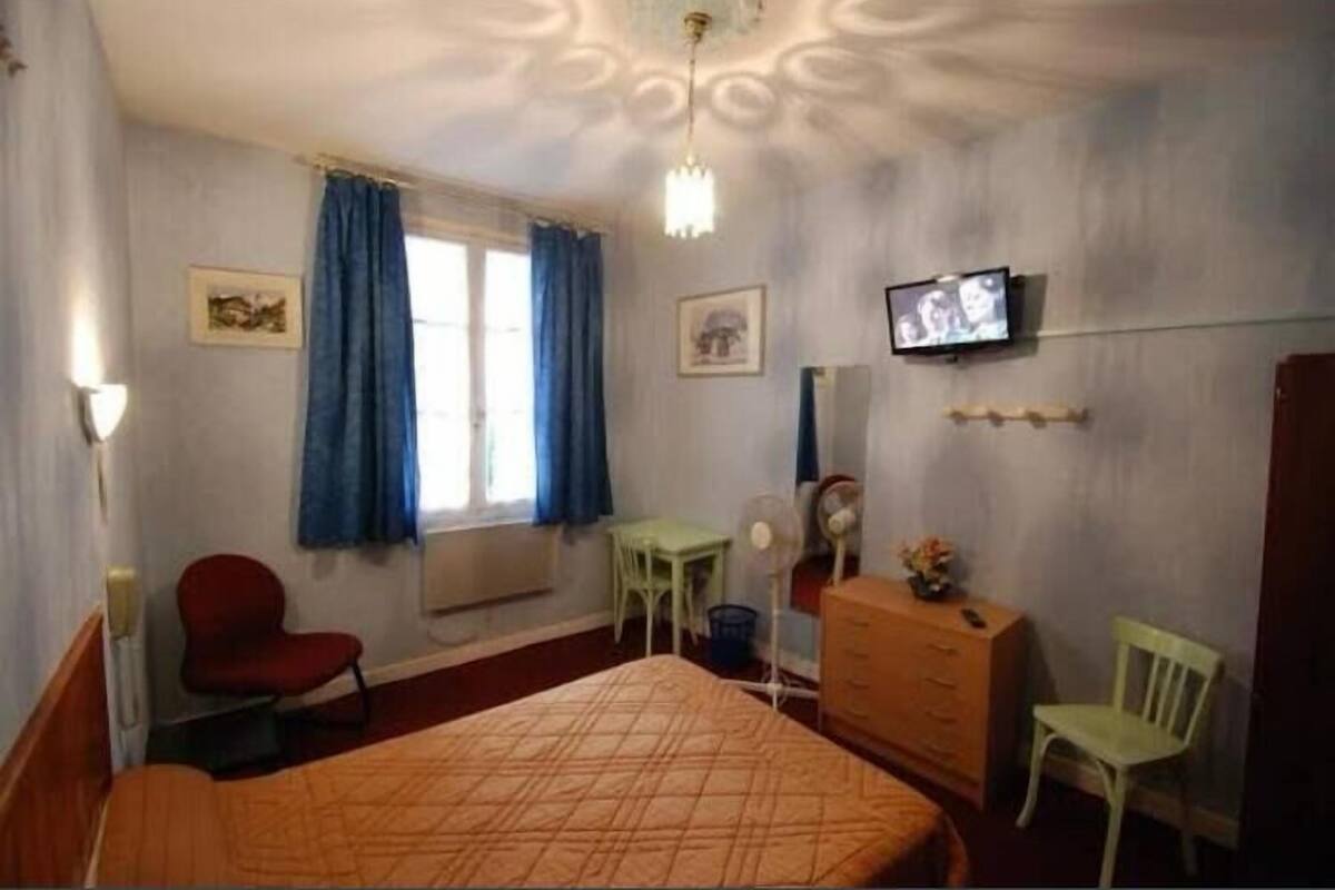Quarto de hotel com cama, escrivaninha, cadeira, cômoda, televisão e janela com cortinas.
