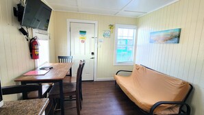 A compact room with a wooden table, chairs, a sofa, a television mounted on the wall, a fire extinguisher, and a window with blinds. - Set Sail! DB6--2 BR/1 Bath Cottage with Kitchen, Walk to the Beach, Dogs OK! (Port Aransas)